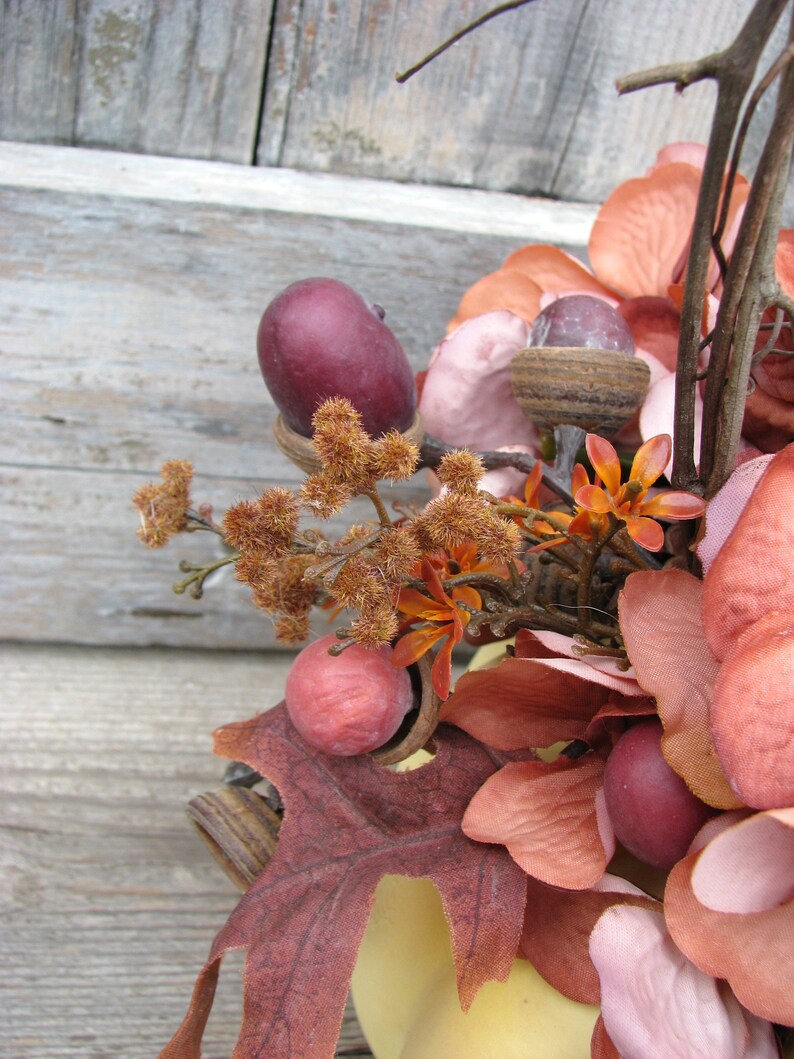 May include: A fall-themed arrangement featuring a variety of faux leaves, flowers, and acorns in shades of brown, pink, and red. The arrangement is displayed in a yellow ceramic container.
