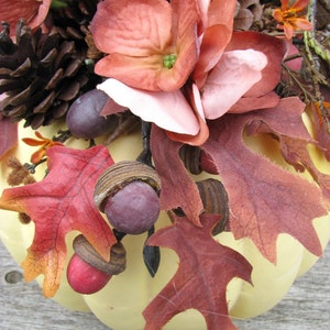 May include: A yellow pumpkin decorated with fall foliage, pinecones, and faux flowers. The pumpkin is decorated with red, brown, and pink flowers, along with brown pinecones and red and brown leaves.