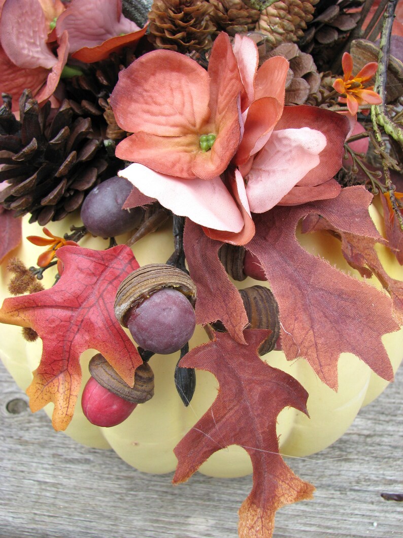 May include: A yellow pumpkin decorated with fall foliage, pinecones, and faux flowers. The pumpkin is decorated with red, brown, and pink flowers, along with brown pinecones and red and brown leaves.