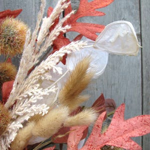 May include: Close-up of a fall-themed arrangement featuring dried plants and leaves. The composition includes reddish-orange oak leaves, fluffy tan grasses, and seed pods, all set against a weathered wooden background. The colors evoke autumn.