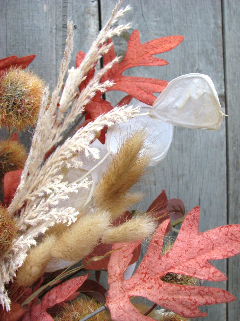 May include: Close-up of a fall-themed arrangement featuring dried plants and leaves. The composition includes reddish-orange oak leaves, fluffy tan grasses, and seed pods, all set against a weathered wooden background. The colors evoke autumn.