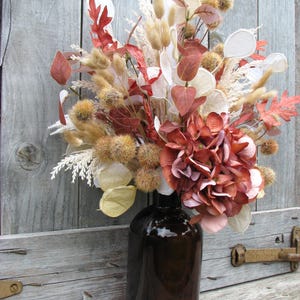 May include: A decorative floral arrangement in a dark brown glass bottle. The bouquet features dried flowers and foliage in shades of red, brown, beige, and white. The arrangement includes hydrangea, seed pods, and various dried grasses, creating a rustic, autumnal aesthetic.