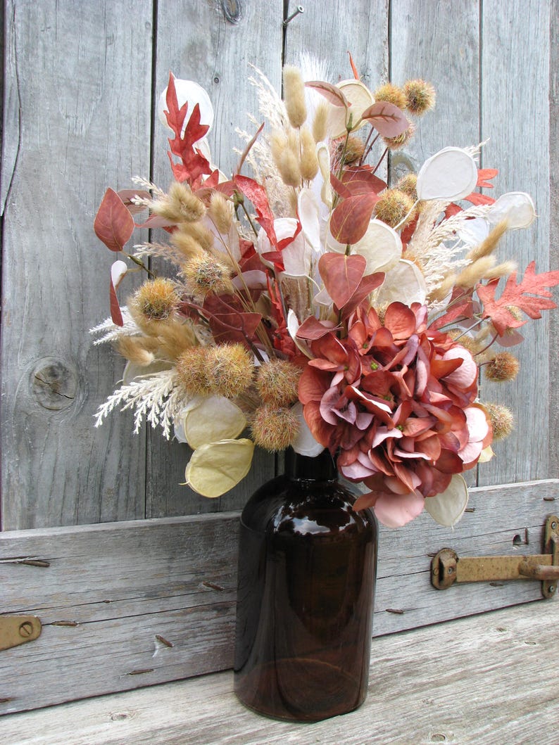 May include: A decorative floral arrangement in a dark brown glass bottle. The bouquet features dried flowers and foliage in shades of red, brown, beige, and white. The arrangement includes hydrangea, seed pods, and various dried grasses, creating a rustic, autumnal aesthetic.