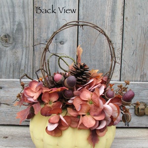 May include: A yellow pumpkin with a wire wreath and fall foliage, including brown acorns, pine cones, and burgundy flowers. The text "Back View" is visible on a wooden background.