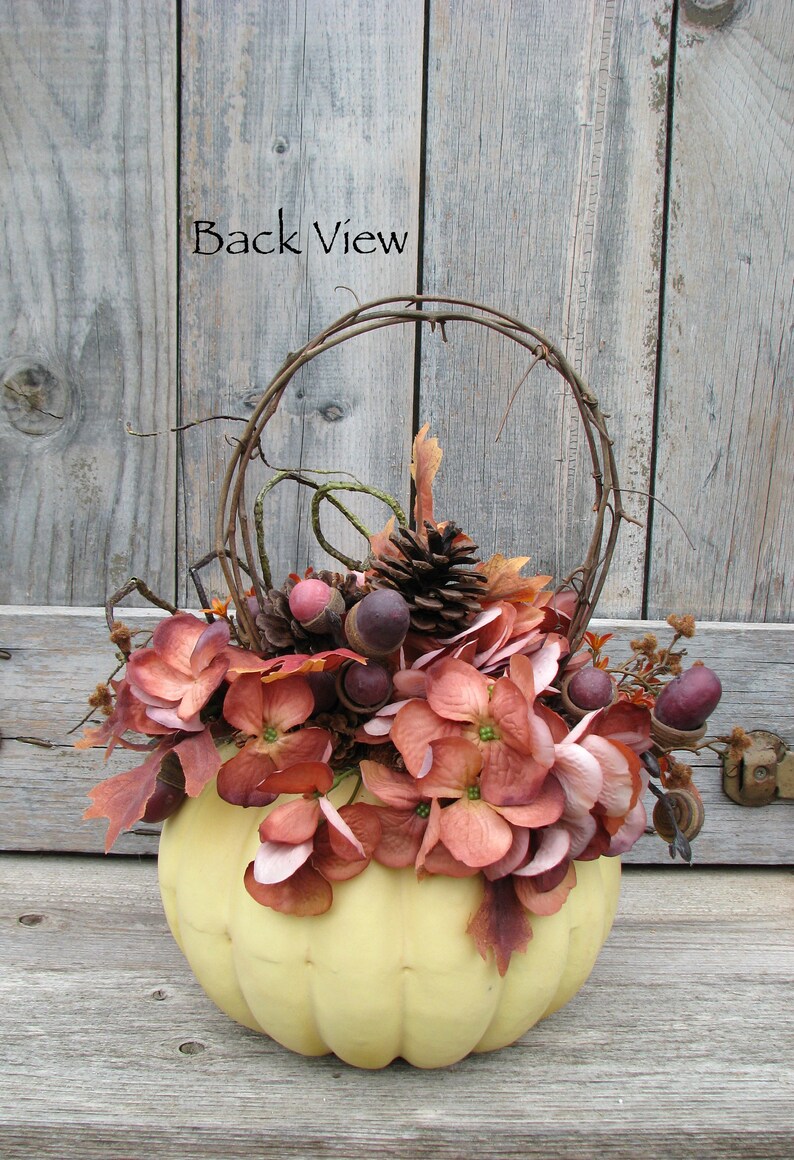 May include: A yellow pumpkin with a wire wreath and fall foliage, including brown acorns, pine cones, and burgundy flowers. The text "Back View" is visible on a wooden background.