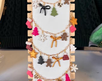 Puede incluir: Cinco cadenas doradas con varios dijes con temática navideña, incluyendo copos de nieve, hombres de jengibre, árboles de Navidad, estrellas y lazos. Las cadenas se muestran en un soporte de madera.