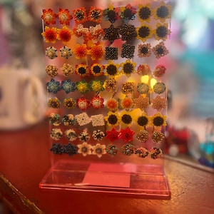Puede incluir: Exhibición de pendientes de botón con cuentas de varios colores y diseños. Los pendientes están dispuestos en un soporte de acrílico transparente, mostrando patrones florales, de estrella y geométricos. Los colores incluyen amarillo, naranja, rojo, azul y negro. Los pendientes están sobre una superficie roja.