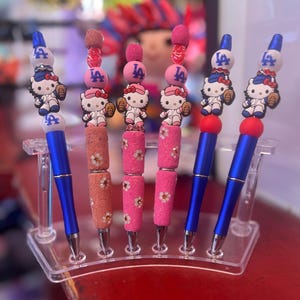以下が含まれることがあります： 青いバレルとピンクのバレルを持つ6本のペン。Hello Kittyのキャラクターが野球のユニフォームを着て、LAのロゴが描かれています。ペンは透明なプラスチックスタンドに飾られています。