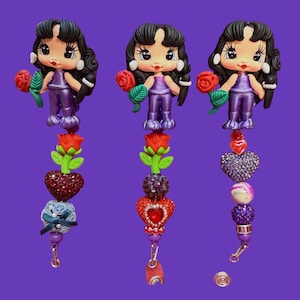 May include: Three decorative doll charms with black hair, holding red roses. The dolls wear purple outfits and are attached to beaded dangles with heart and floral accents. The background is a solid purple.