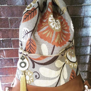 Könnte beinhalten: Ein beiger Rucksack mit Kordelzug und einem Blumenmuster in Braun-, Orange- und Cremetönen. Der Rucksack hat eine Reißverschlusstasche und einen braunen Lederboden.