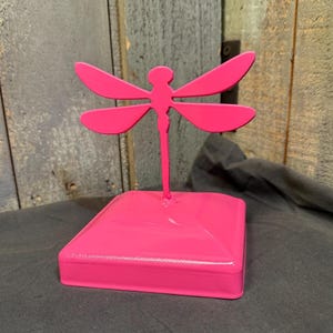 May include: A bright pink metal dragonfly sculpture mounted on a square base. The dragonfly features four symmetrical wings extending from a central body, all finished in a solid, glossy pink color. The entire object is constructed from metal.