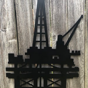 May include: Black metal silhouette of an offshore oil rig with a tall derrick and a crane arm. The silhouette is mounted on a wooden fence.