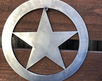 May include: A silver metal star within a circle, fixed to a wooden surface. The star is a five-pointed design, and the circle surrounds it. A single screw secures the metal piece to the wood. The wood has a brown, textured appearance.