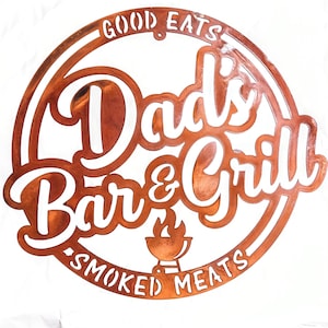 May include: A rusty brown metal sign with white lettering that reads "Good Eats Dad's Bar & Grill Smoked Meats". The sign has a circle shape with a double border and a grill icon in the center.