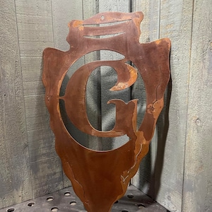 May include: A rusty metal arrowhead shaped wall decor with a cut-out letter "G" in the center.