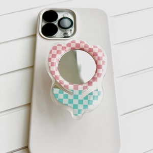 May include: A white phone case with a pink and blue checkered phone ring holder with a mirror in the center.