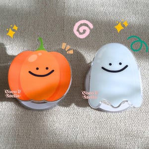 May include: Two Halloween-themed phone grips. One is an orange pumpkin with a green stem and a smiling face. The other is a white ghost with a smiling face. Both have a white base and are on a neutral background.