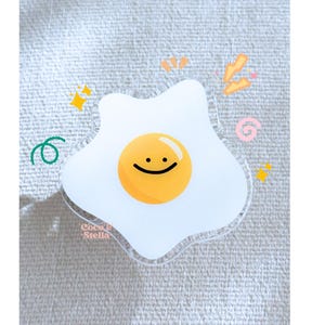 May include: A clear acrylic sticker in the shape of a fried egg with a smiling face. The yolk is yellow, and the white is translucent. Decorative elements include stars, swirls, and the text "Coco & Stella".