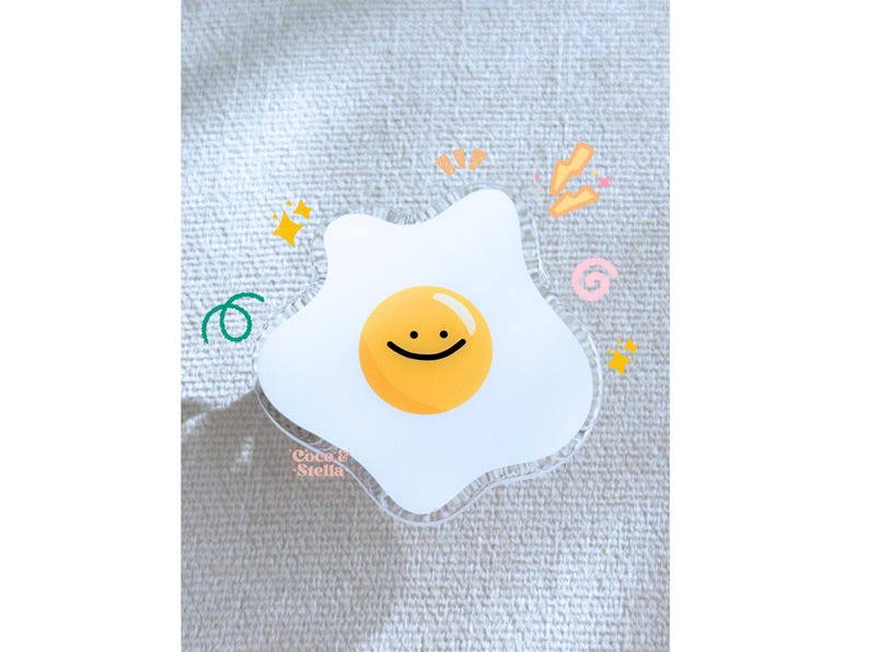 May include: A clear acrylic sticker in the shape of a fried egg with a smiling face. The yolk is yellow, and the white is translucent. Decorative elements include stars, swirls, and the text "Coco & Stella".