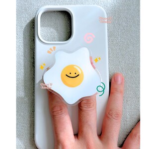 May include: White phone case with a fried egg design. The egg has a yellow yolk with a smiling face and a white egg white. The case has the text "Coco & Stella" and other decorative elements.