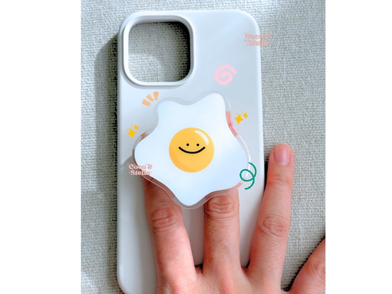 May include: White phone case with a fried egg design. The egg has a yellow yolk with a smiling face and a white egg white. The case has the text "Coco & Stella" and other decorative elements.