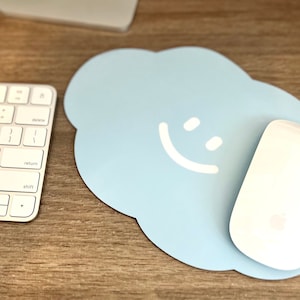 May include: A light blue cloud-shaped mousepad with a white smiley face design. A white wireless mouse is on the mousepad.