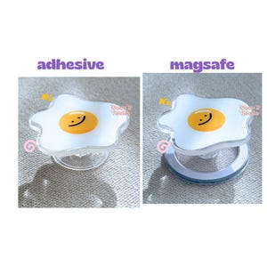 May include: Clear phone grip with a white, egg-shaped design and a yellow yolk with a smiling face. The text "adhesive" and "magsafe" are above the grips. The brand name "Coco & Stella" is also visible.