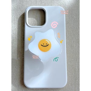 May include: A light gray phone case featuring a cheerful fried egg design with a smiling face. The case has a cutout for the camera and is decorated with colorful doodles. The text "Coco Stella" is visible.