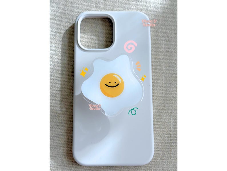 May include: A light gray phone case featuring a cheerful fried egg design with a smiling face. The case has a cutout for the camera and is decorated with colorful doodles. The text "Coco Stella" is visible.