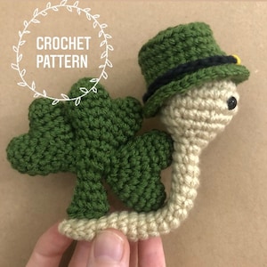 Puede incluir: Patrón de crochet para un trébol verde con un duende beige que lleva un sombrero verde con una banda negra y un botón dorado.