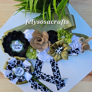 May include: A green, black, and white sash with a cow print design. The sash has a large flower arrangement with black, white, green, and brown fabric flowers. The sash also has two smaller pins with the text "Mommy to Be" and "Daddy to Be".