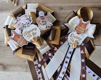Puede incluir: Dos cintas de baby shower con temática de café, con las frases "Mommy To Be" y "Daddy To Be". Las cintas son marrones, beige y blancas, decoradas con motivos de granos y tazas de café. Se ve una guirnalda de cuentas de madera.