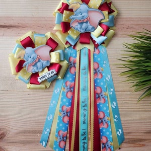 May include: Two blue, yellow, and red baby shower sashes with a Dumbo the Elephant design. One sash says "Daddy to Be" and the other says "Mommy to Be! Mommy to Be! Mommy to Be!"