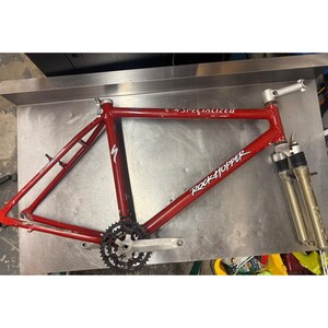 Puede incluir: Cuadro de bicicleta de monta&ntilde;a Specialized Rockhopper rojo con juego de bielas y horquilla delantera. El cuadro tiene las palabras "SPECIALIZED" y "ROCKHOPPER" impresas. El cuadro de la bicicleta est&aacute; sobre una superficie met&aacute;lica.