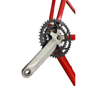 Puede incluir: Primer plano de un juego de bielas de bicicleta con una biela plateada y un plato de cadena oxidado y oscuro. El juego de bielas est&aacute; fijado a un cuadro de bicicleta rojo, lo que indica un estado vintage o usado. Fondo blanco.