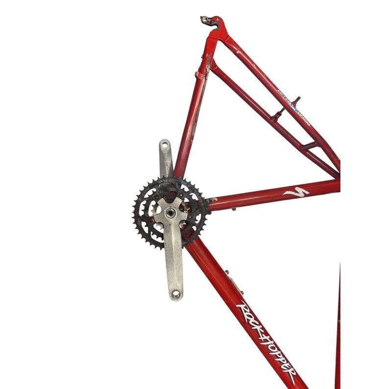 Puede incluir: Un cuadro de bicicleta rojo con un plato y biela negros. El cuadro tiene la inscripci&oacute;n "Rock Hopper". El cuadro de la bicicleta est&aacute; sobre un fondo blanco.