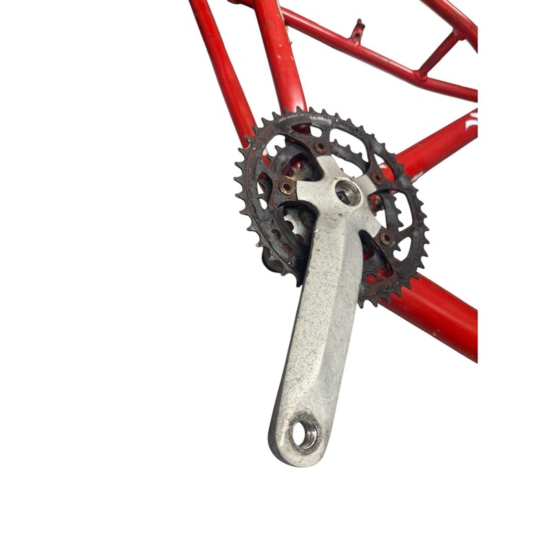 Puede incluir: Primer plano de un juego de bielas de bicicleta con un brazo de pedal plateado y un plato de cadena oxidado, gris oscuro. El juego de bielas est&aacute; fijado a un cuadro de bicicleta rojo, mostrando los componentes mec&aacute;nicos.