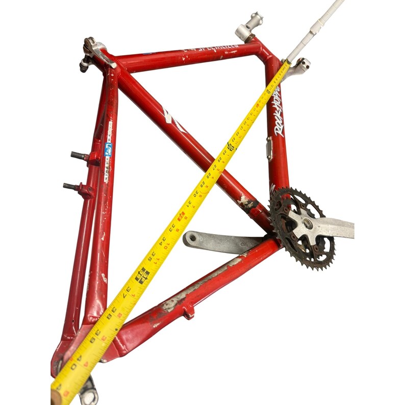 Puede incluir: Un cuadro de bicicleta rojo con una cinta m&eacute;trica amarilla colocada sobre &eacute;l. El cuadro tiene un plato y un juego de bielas adjuntos. La cinta m&eacute;trica muestra una longitud de aproximadamente 101 cm.