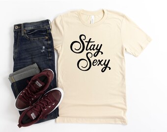 Op de afbeelding: Een beige t-shirt met de tekst "Stay Sexy" in zwarte cursieve letters. Het shirt wordt gecombineerd met donkerblauwe jeans en bordeauxrode high-top sneakers.