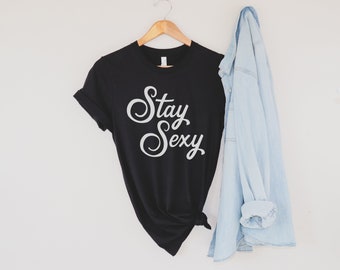 Op de afbeelding: Een zwart T-shirt met de tekst "Stay Sexy" in witte cursieve letters. Het shirt is gevouwen en hangt aan een houten hanger. Een lichtblauw denim jack is over de hanger gehangen.