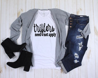 May include: A casual outfit laid out on a light wood surface. The items include a white t-shirt with the text "triflers need not apply", a grey cardigan, distressed blue jeans, and black ankle boots.