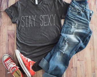 Op de afbeelding: Een donkergrijs T-shirt met de tekst "STAY SEXY" in witte omrande letters. Het T-shirt wordt gecombineerd met een blauwe jeans en rode sneakers.