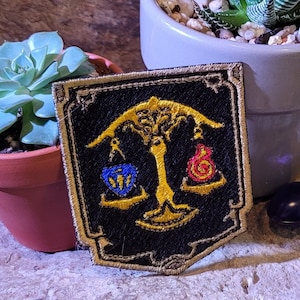 Pode incluir: Um remendo bordado preto e dourado com uma árvore, uma balança e um símbolo azul e vermelho. O remendo está sobre uma superfície de madeira com três suculentas em vasos.