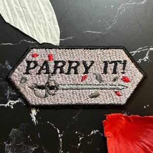 Pode incluir: Patch bordado cinza com as palavras "PARRY IT!" e um desenho de espada. O patch tem detalhes em vermelho e cinza. Pétalas vermelhas e brancas estão espalhadas ao redor do patch em uma superfície de mármore preto.