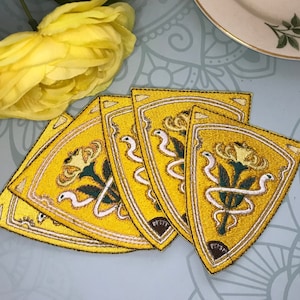May include: Five yellow embroidered iron-on patches in the shape of a shield with a stylized white flower and green stem inside. The flower is surrounded by a white ribbon with a white snake wrapped around it.