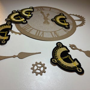 Puede incluir: Un conjunto de cinco parches de esfera de reloj negros y dorados con costuras amarillas. Los parches están dispuestos alrededor de una esfera de reloj de papel marrón con números romanos. También hay dos manecillas de reloj de papel marrón y un engranaje de papel marrón.