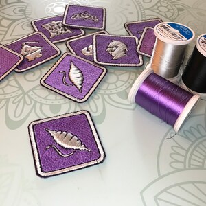 May include: Purple felt patches with silver embroidery of various designs, including a pod, a crown, and a stick. Three spools of thread in silver, black, and purple.