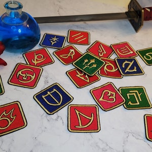 May include: A collection of 17 colourful square patches with gold embroidered symbols. The patches are scattered on a white marble surface. A blue glass bottle and a red and black katana sword are also visible in the image.