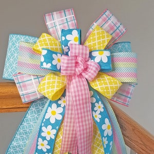 May include: A colorful bow made with various patterned ribbons, including yellow with white daisies, pink gingham, blue with white daisies, and green and pink chevron. The bow is tied around a wooden railing.