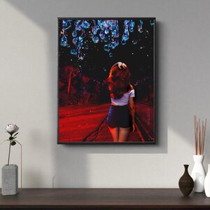 May include: A framed art print featuring a person walking away on a red-lit road under a dark sky with blue and white droplets. The person is wearing a white top and dark shorts. The artwork is displayed on a wall above a wooden table.